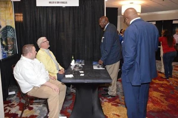 Several people converse around a table at a conference booth in a carpeted convention hall.