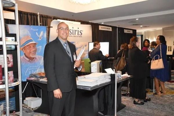 Conference booth with attendees, a man in a black suit, and promotional banners at an indoor event.