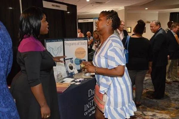 Two women converse at a conference booth in a busy hotel lobby.