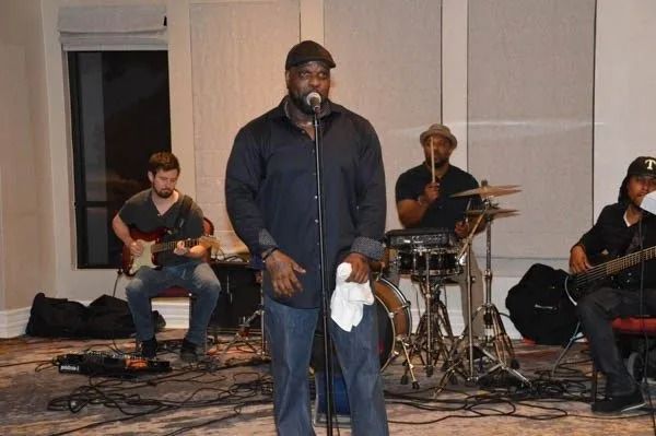 Musician singing into a microphone with a band playing behind him on a small indoor stage