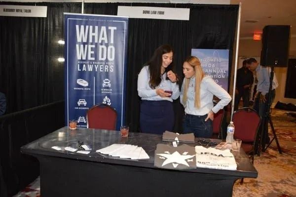 Law firm booth with “WHAT WE DO” banner, brochures on table, and two women talking at a trade show.