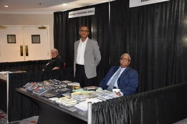 Three people at a table covered with books and pamphlets in a booth with black curtains.