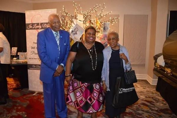 Three people posing indoors at an event, with one holding a bag and a decorated backdrop behind them.