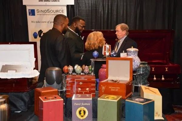 Three men converse at a display table of colorful engraved urns and boxes at a trade show booth.
