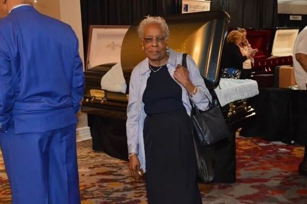 Person standing indoors beside a piano, wearing a dark dress and light jacket, holding a bag.
