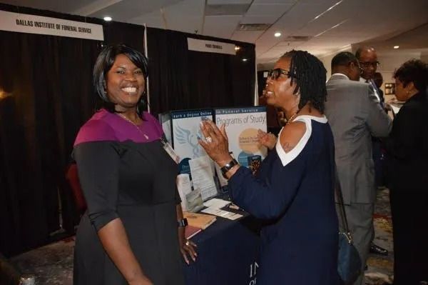 Two women chatting at a booth during an indoor event, one smiling and holding a pamphlet.