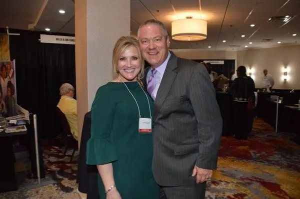 Two smiling people pose together at a conference in a hotel lobby.