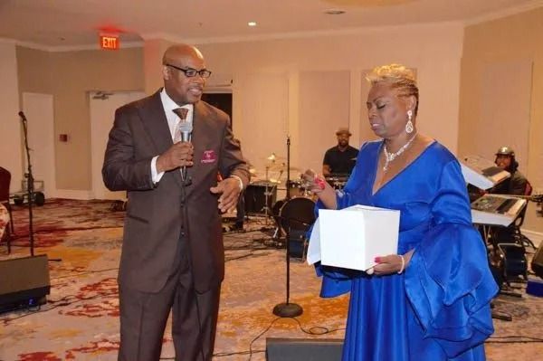 Man in a suit speaking into a microphone beside a woman holding a white box at a formal indoor event