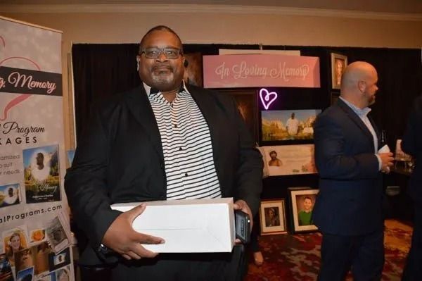 Man holding a white box at an indoor event with display tables and banners in the background