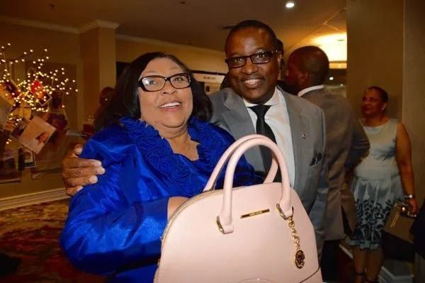 Smiling couple at an indoor event, woman in blue holding a pink handbag, man in a gray suit beside her.