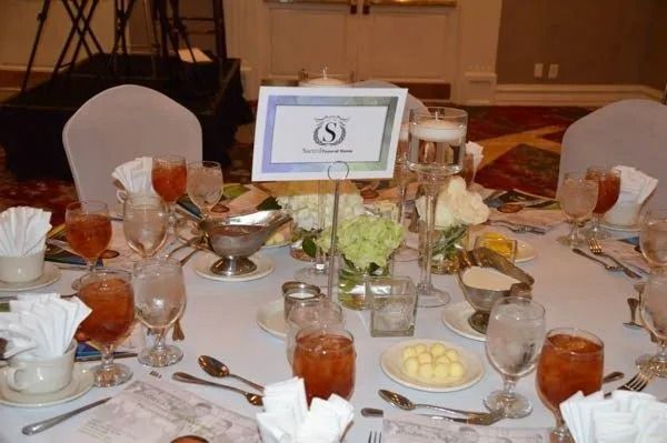 Formal banquet table with white chairs, place settings, glasses, and floral centerpiece in a ballroom