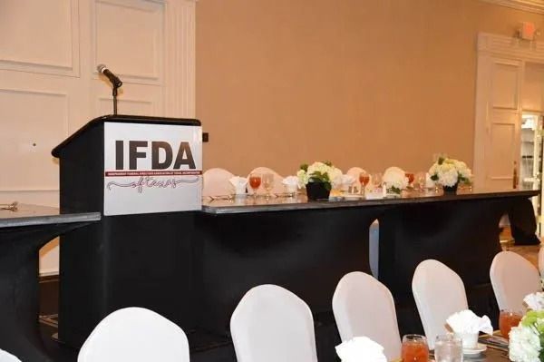 IFDA podium and long black-draped table with white chairs and floral centerpieces in a banquet hall