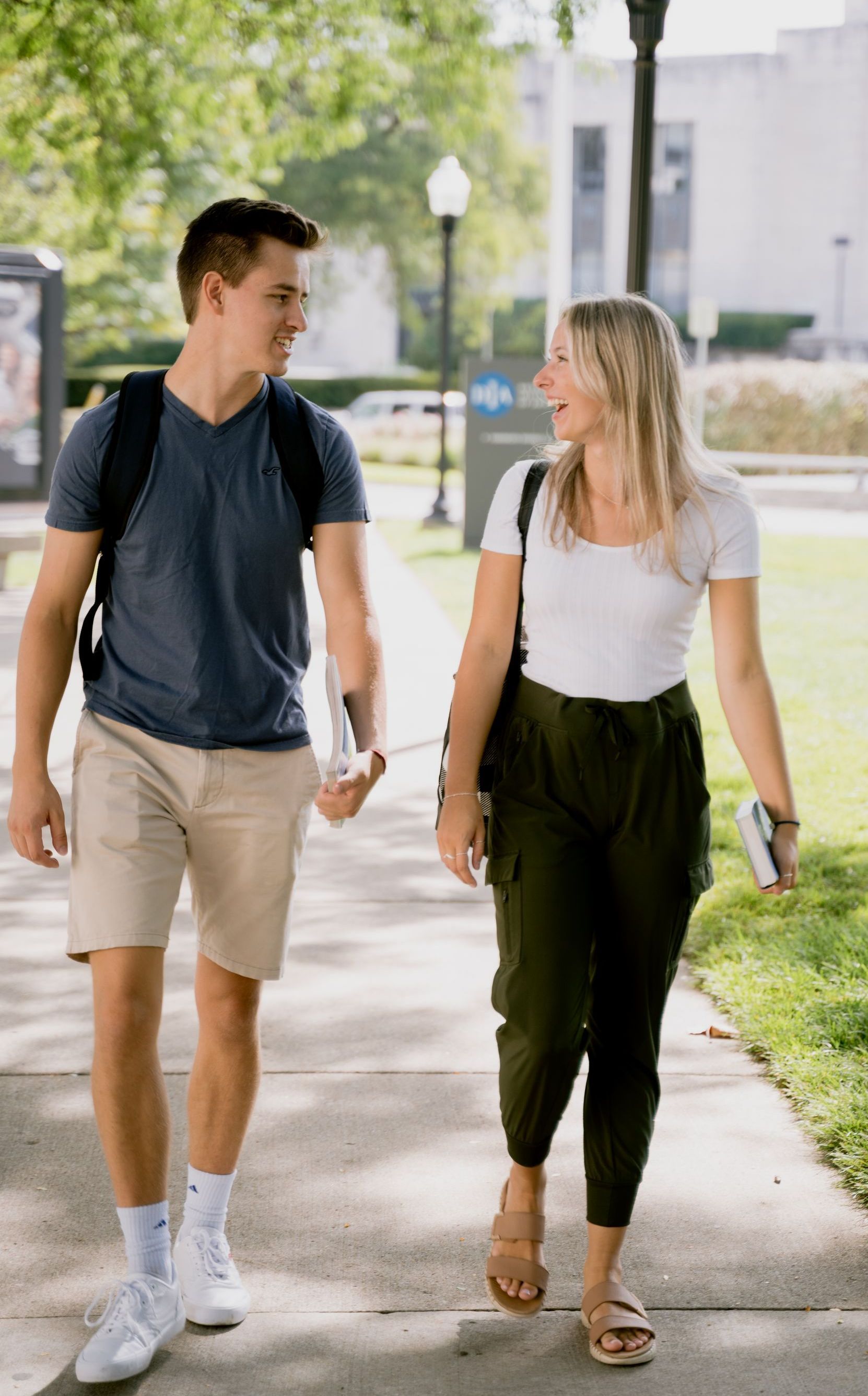 Two students walking and talking on a sunny sidewalk, carrying backpacks.