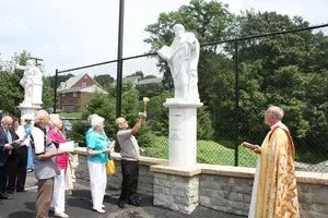 People gathered outdoors around a statue while a robed person leads a blessing or prayer.