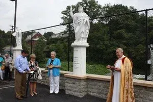 Outdoor religious ceremony beside a white statue, with people gathered on a wet pavement and a priest reading from a book