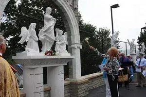White angel statue under an arch with people gathered nearby at an outdoor memorial.