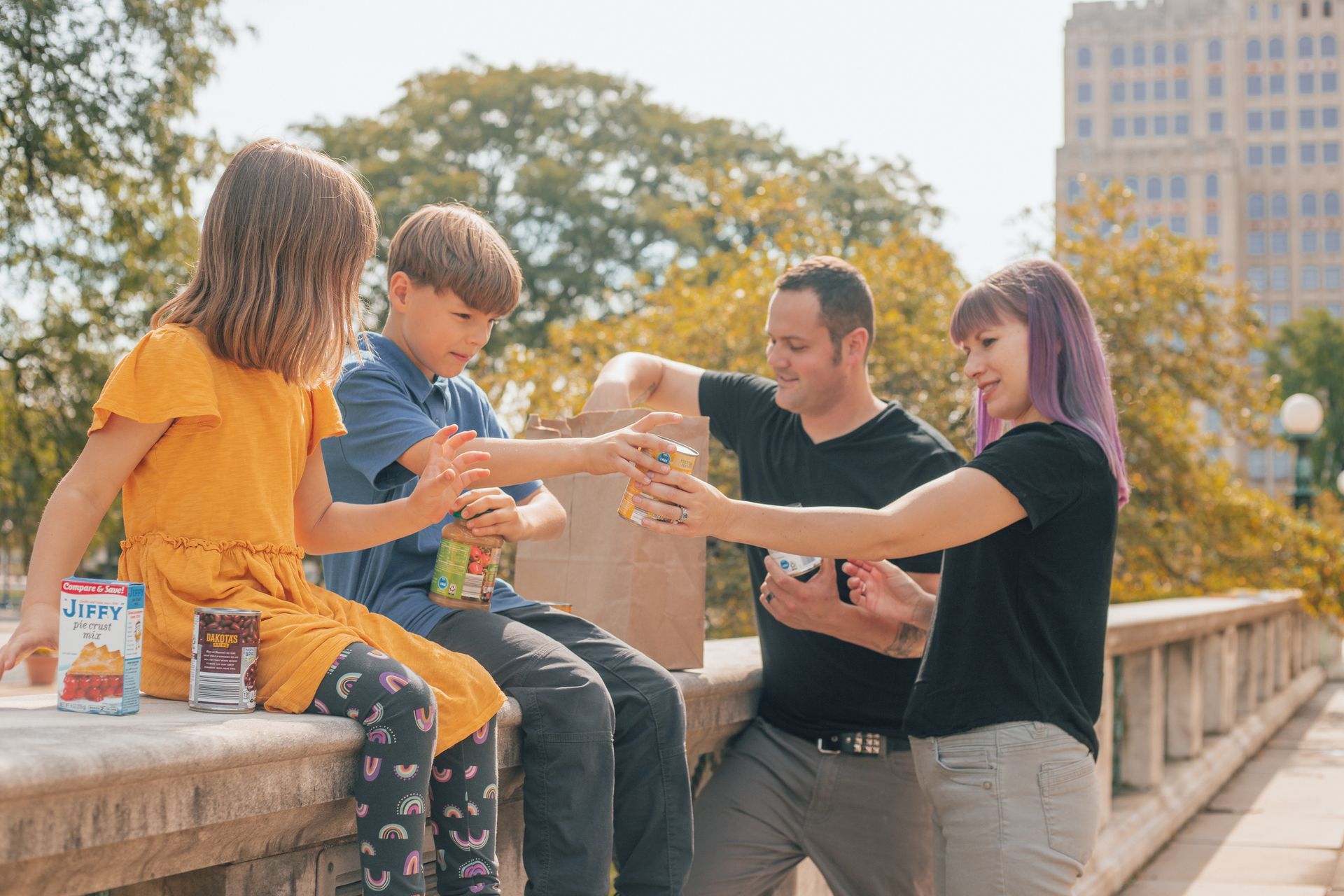 Four friends sharing snacks on a sunny outdoor bench, smiling and passing food together.