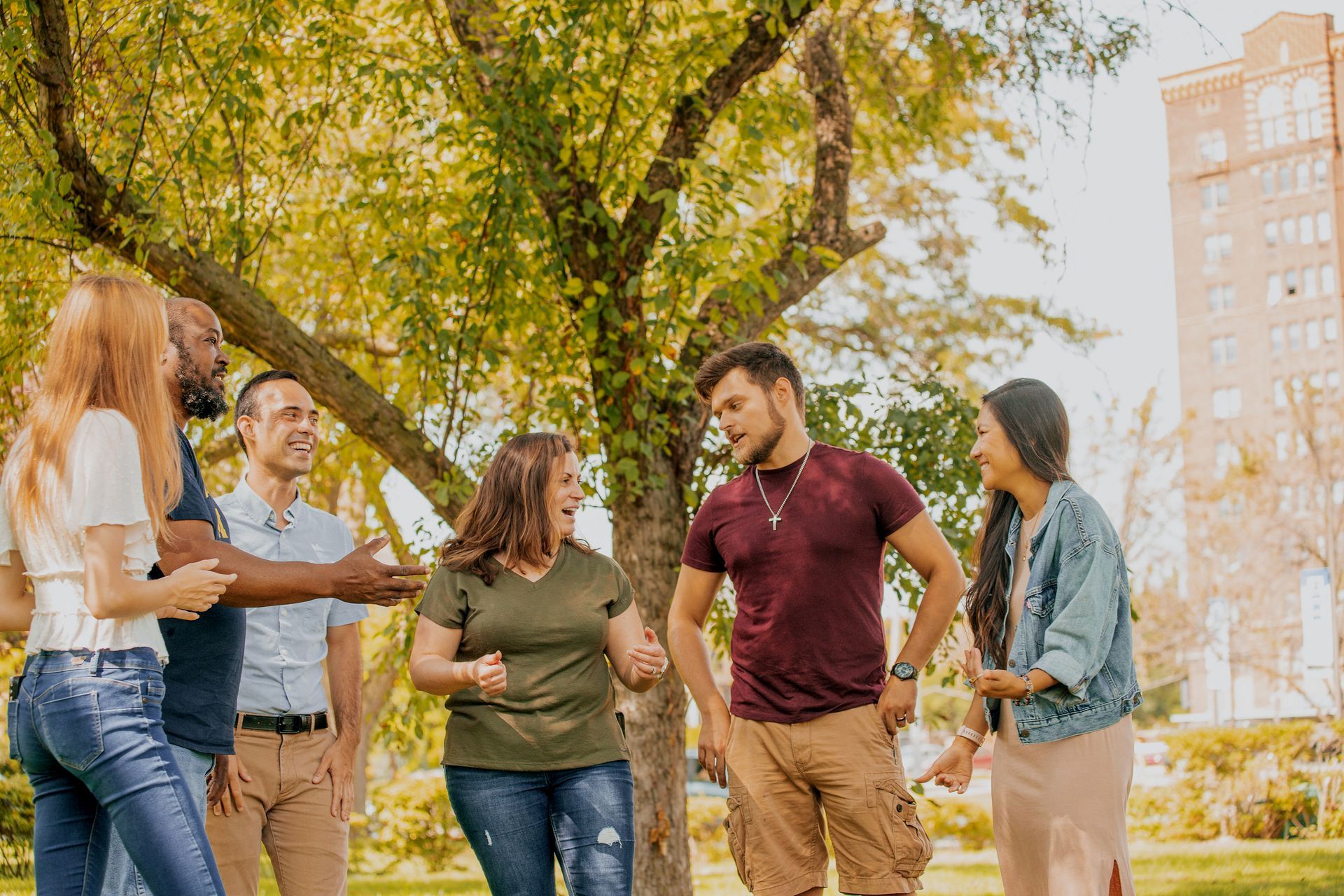 Five friends chatting in a sunny park under trees with city buildings in the background