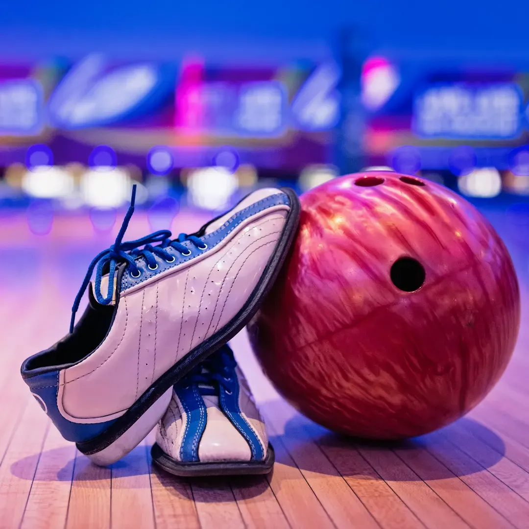 White bowling shoe beside a red bowling ball on a lane, with blurred pins and neon-lit alley background