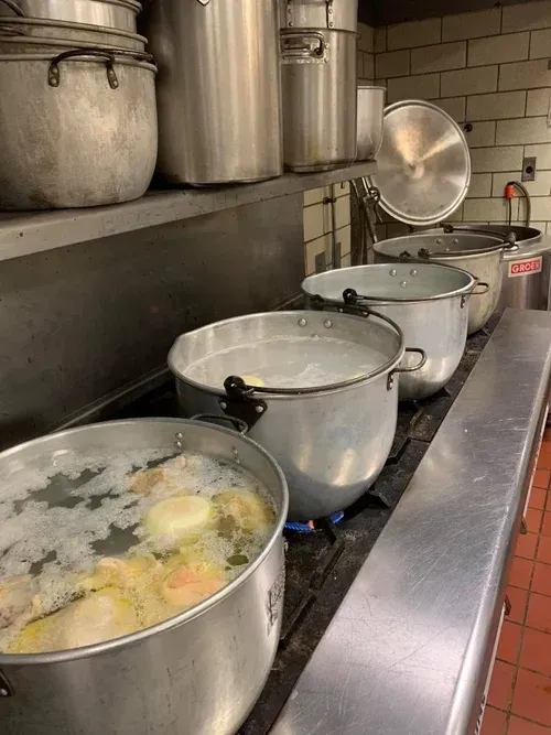 Large pots of food simmering on a commercial kitchen stove with lids and utensils nearby.