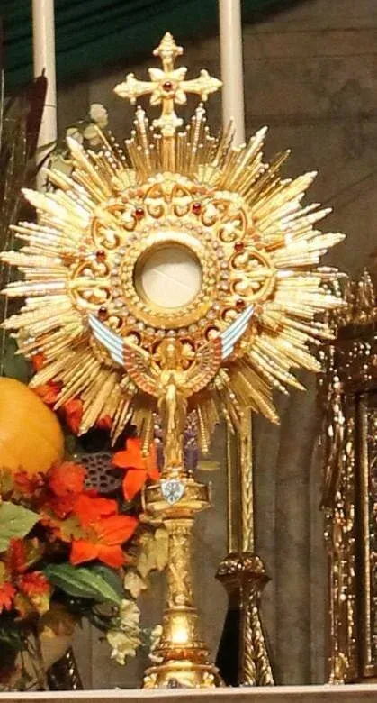 Gold Catholic monstrance with a white host, surrounded by candles and flowers on an altar