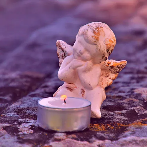 Small angel figurine beside a lit tealight candle on a purple textured surface.