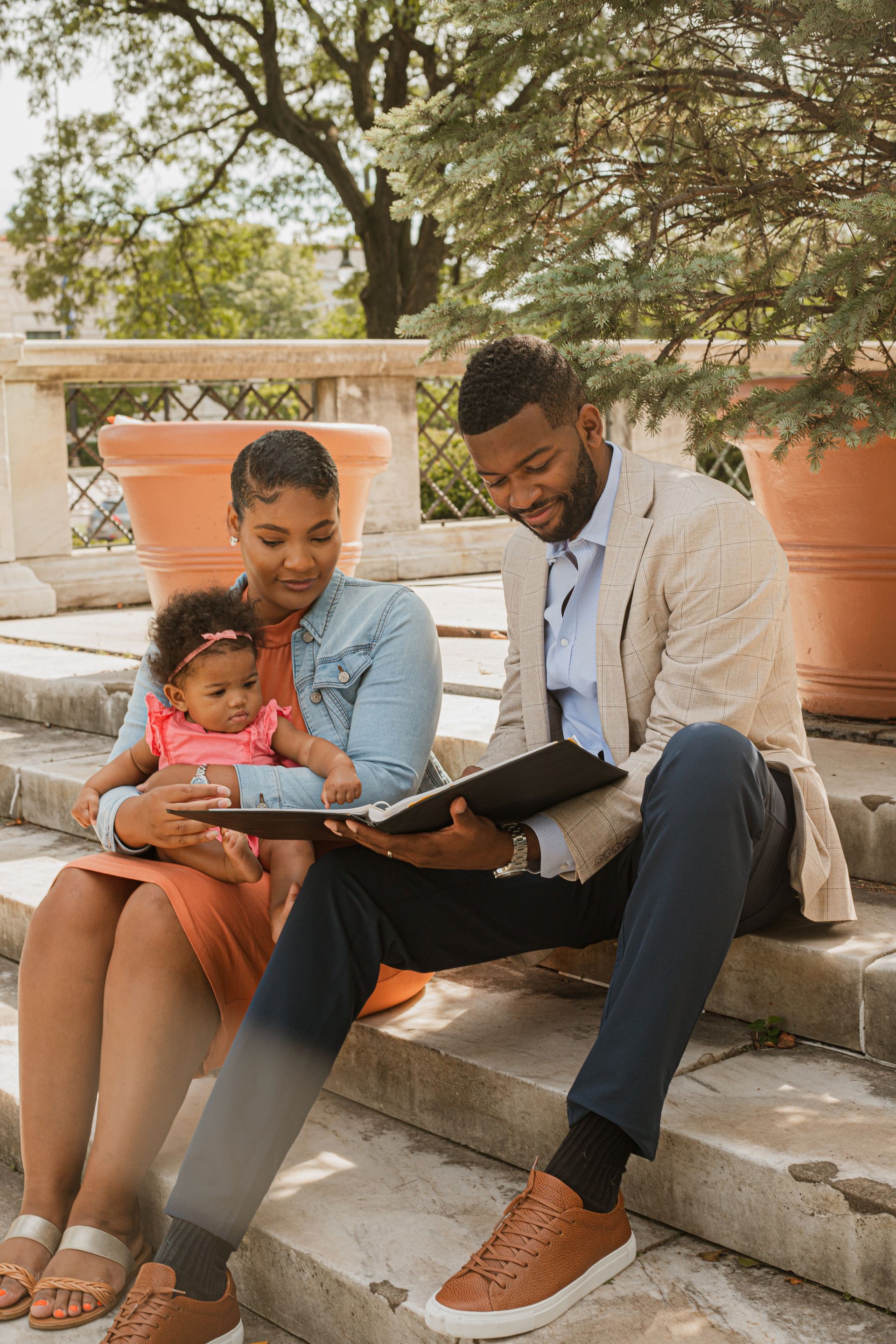 Two adults and a toddler sit on stone steps, looking at a tablet outdoors under trees.