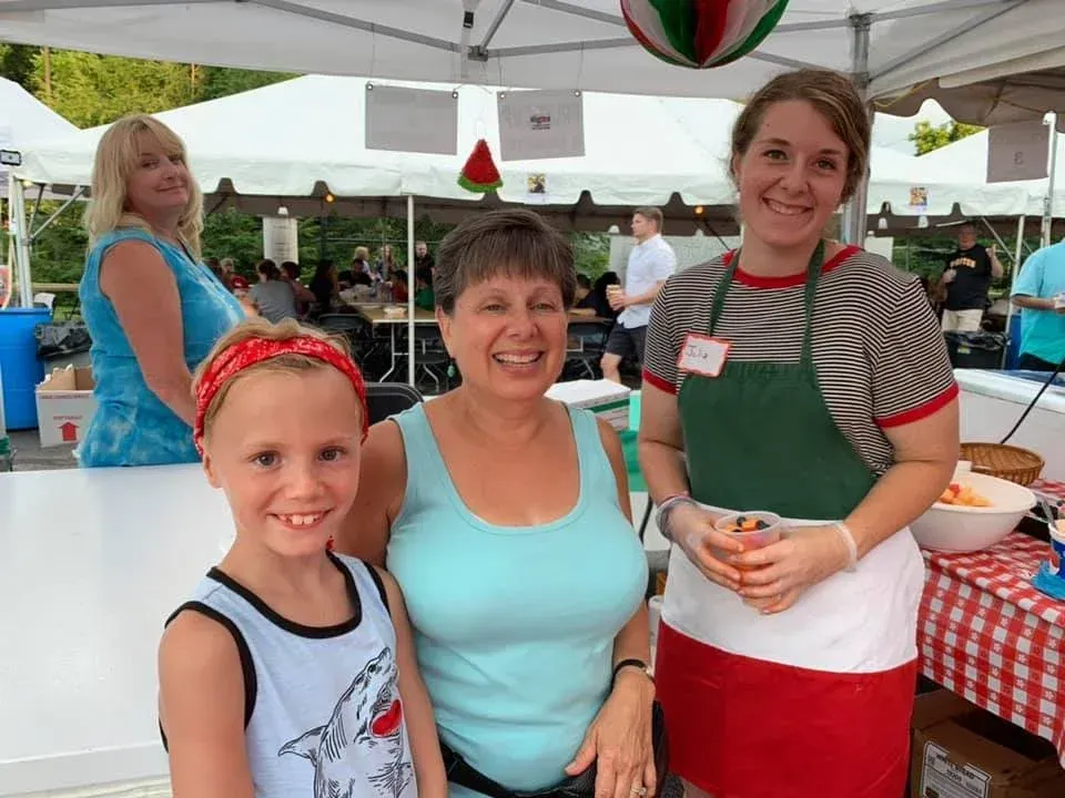Three people smiling at an outdoor market stall under a white canopy, with a child in the foreground