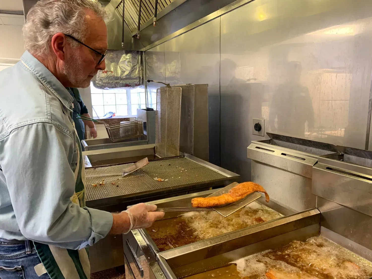 Worker in a kitchen inspecting food on a fryer line under stainless steel ventilation hoods