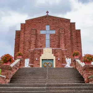 Brick church facade with large cross above a stairway, flanked by flower planters and white statues