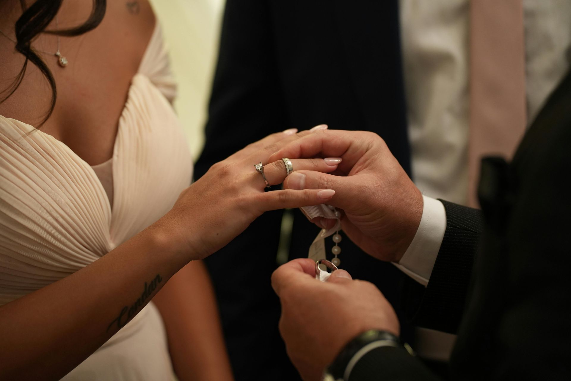 Hands exchange rings during a wedding ceremony, with the bride in a cream dress visible nearby.