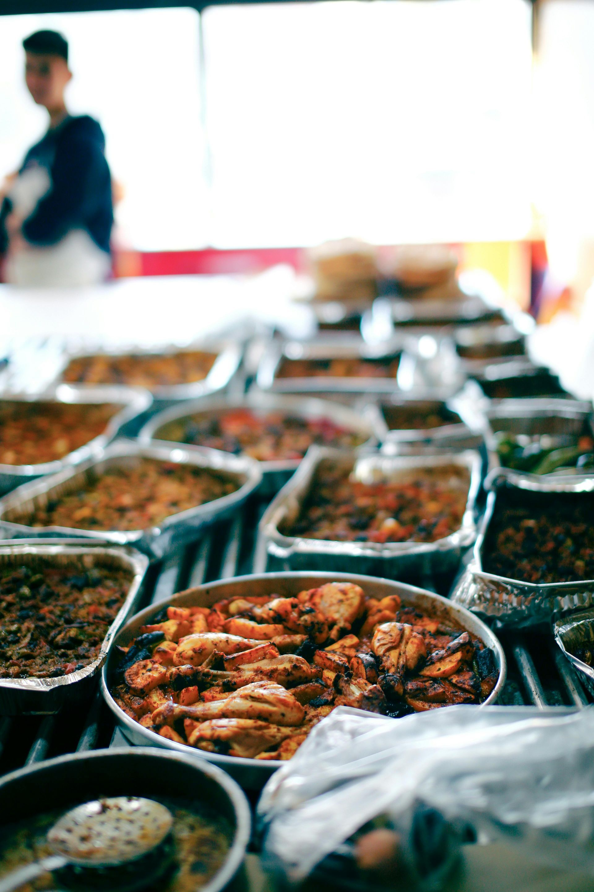 Rows of covered food trays at a market stall, with a bowl of grilled shrimp in the foreground.