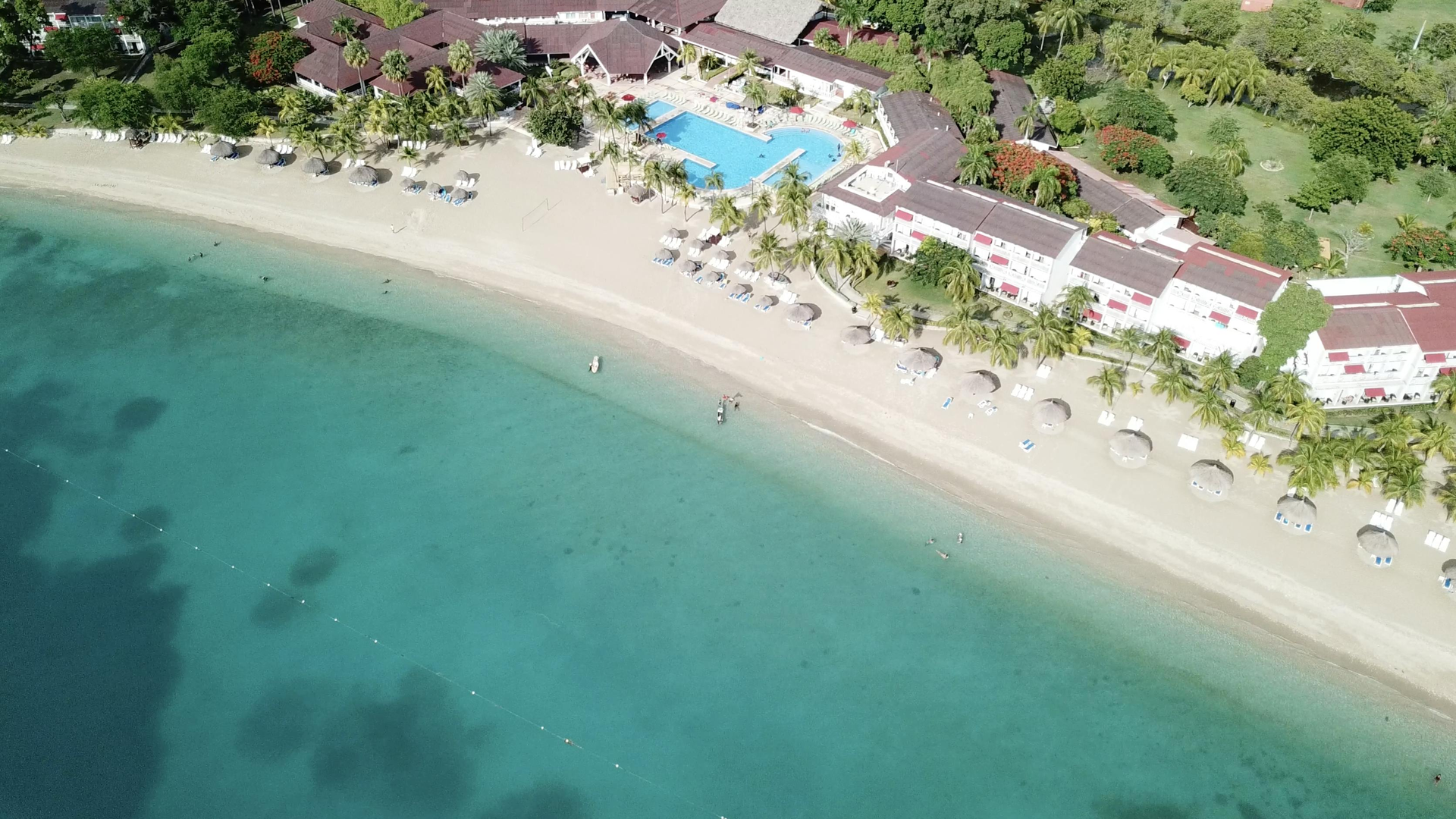 Aerial view of a tropical beach resort with turquoise water, white sand, umbrellas, and a pool near the shore