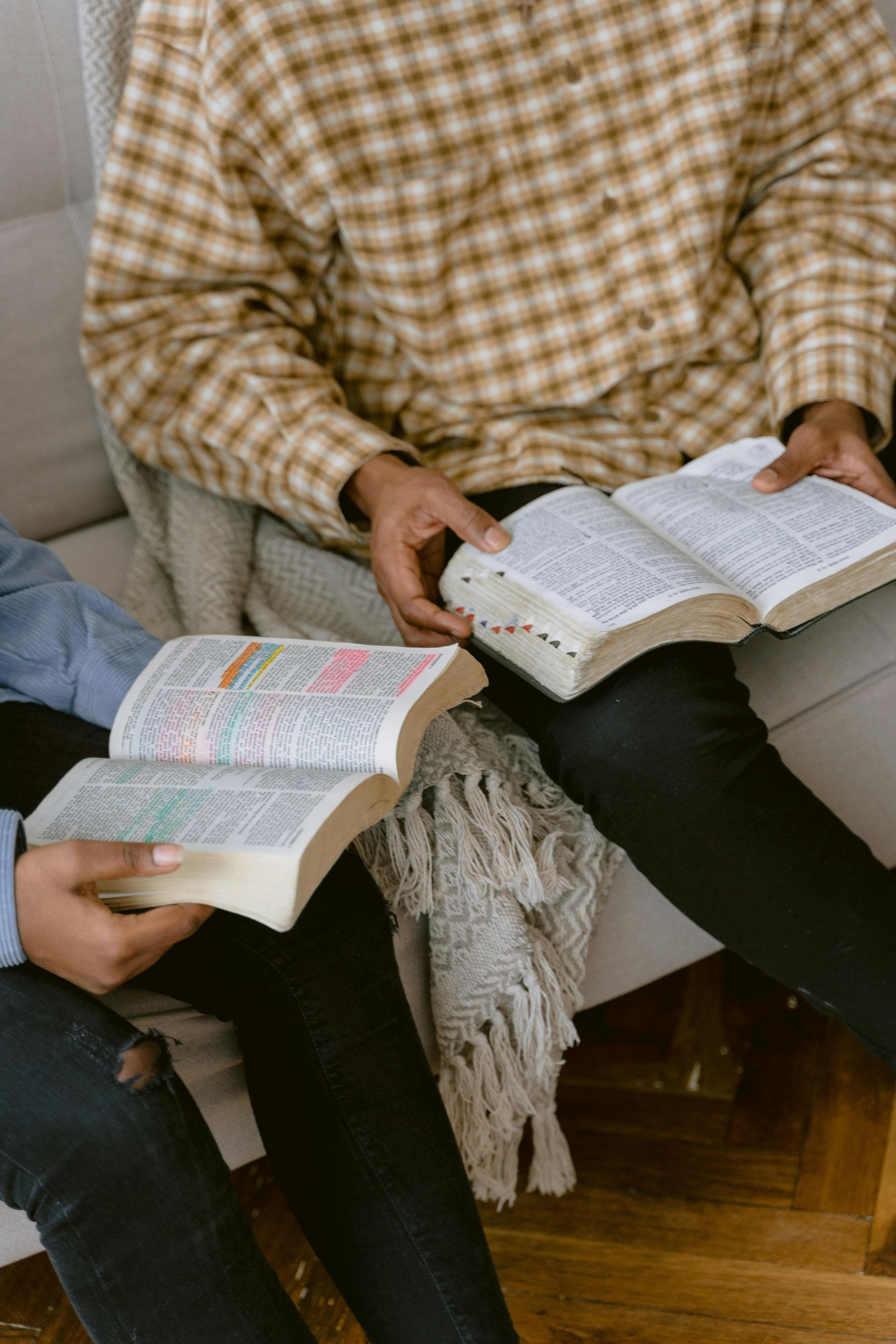 Two people sitting on a couch reading open books, one with a patterned sweater and one with a white notebook.