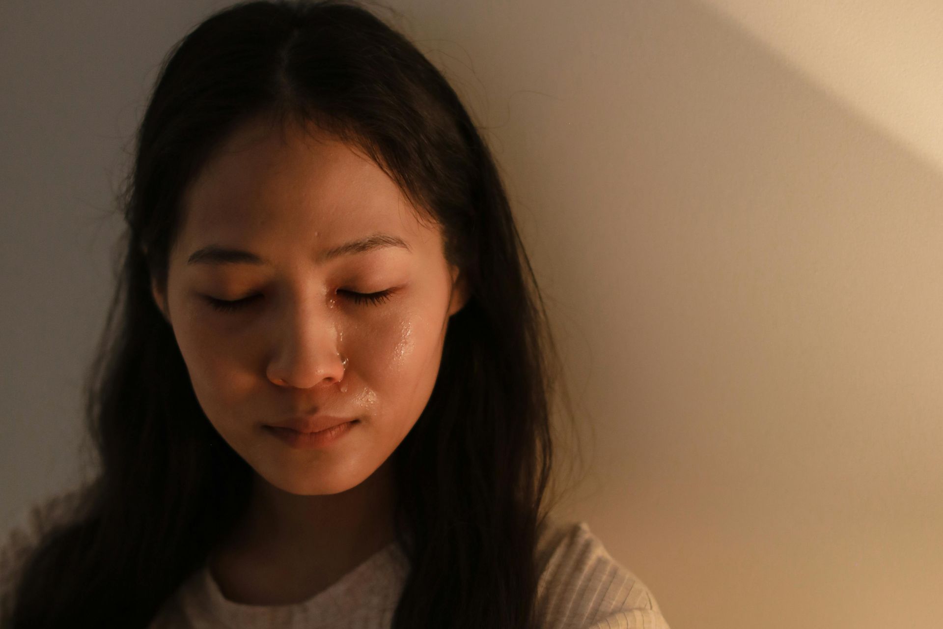 Person with eyes closed in warm, dim light, standing against a plain wall.