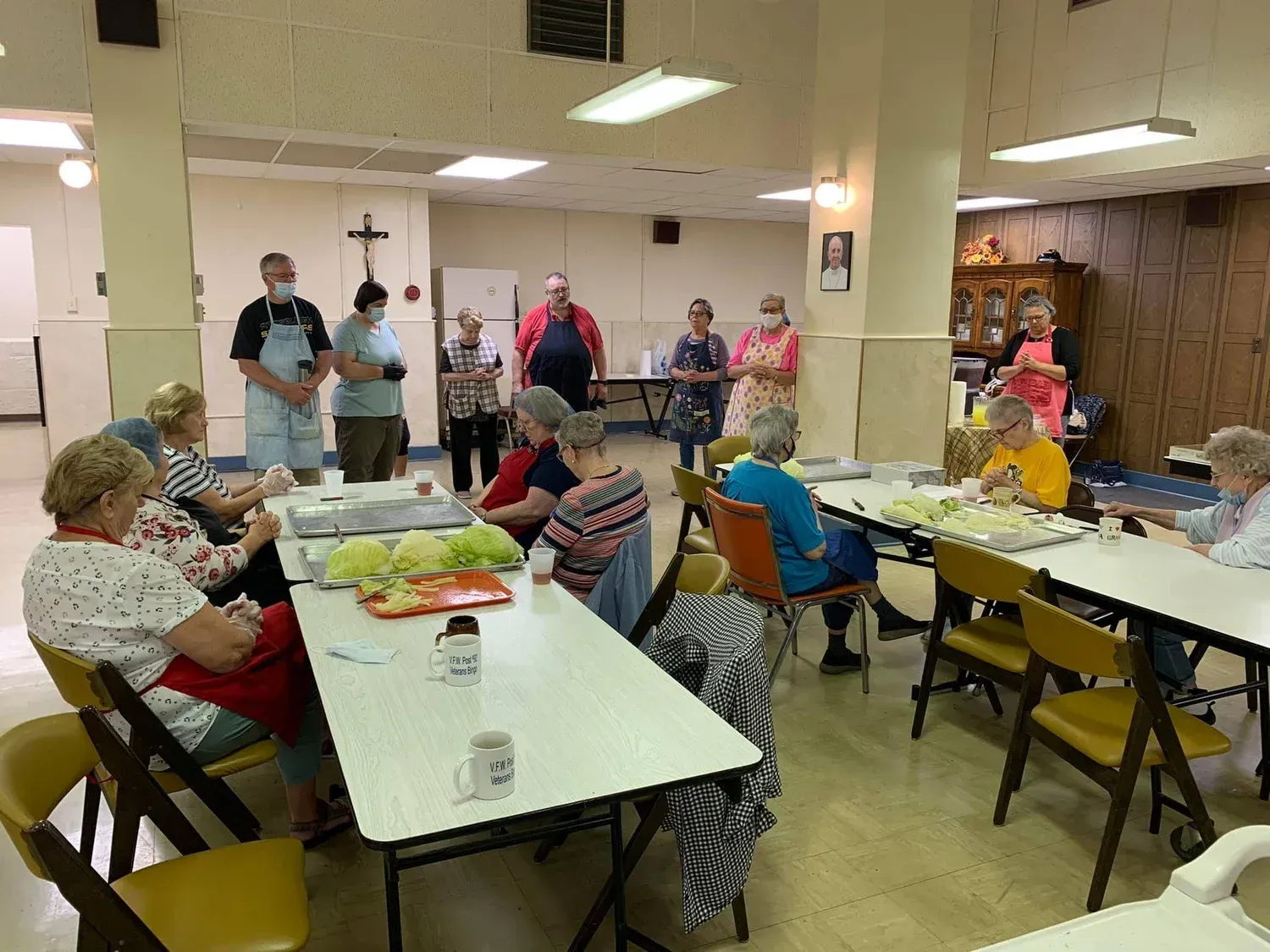 People seated at long tables in a bright community dining hall, with a few standing near the back wall.