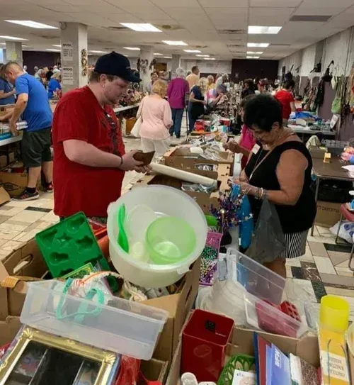 People browsing items at a crowded indoor flea market table with bowls, bins, and assorted goods