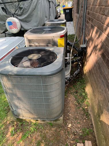 Three outdoor HVAC condenser units beside a brick wall and narrow grass strip with debris nearby
