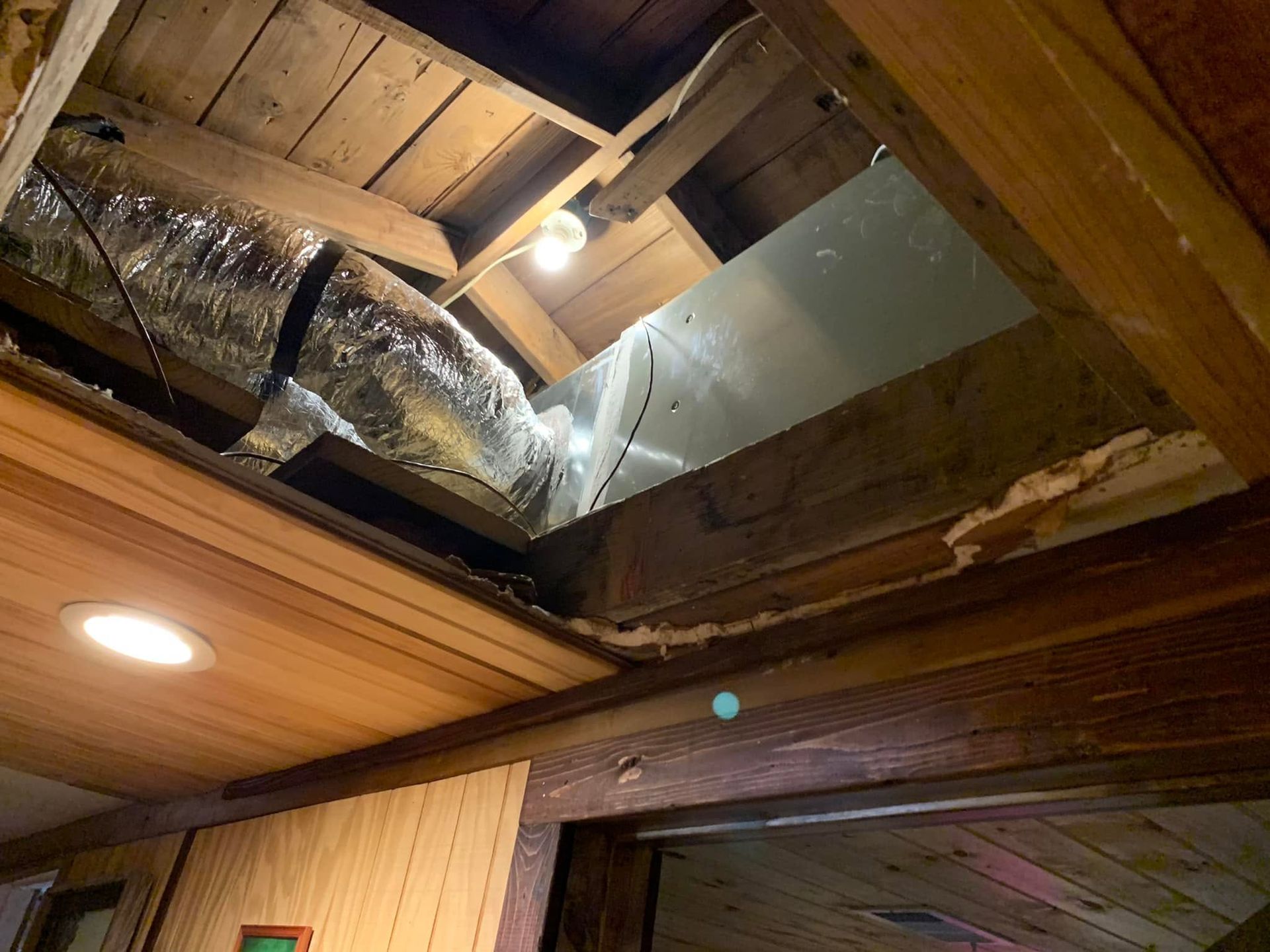 Damaged ceiling with exposed insulation and ducts above a wood-paneled room, lit by a ceiling light