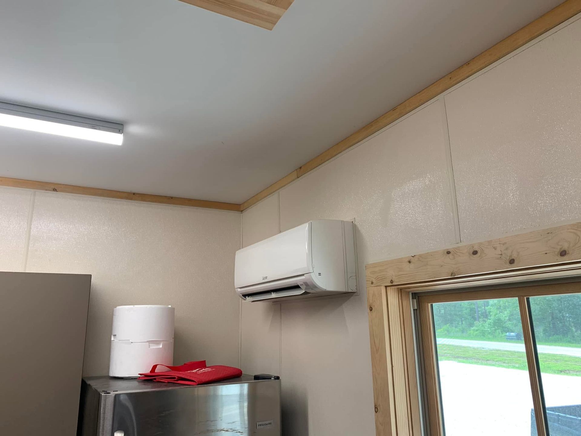 Small room with a wall-mounted air conditioner, white water heater, and refrigerator near a window.