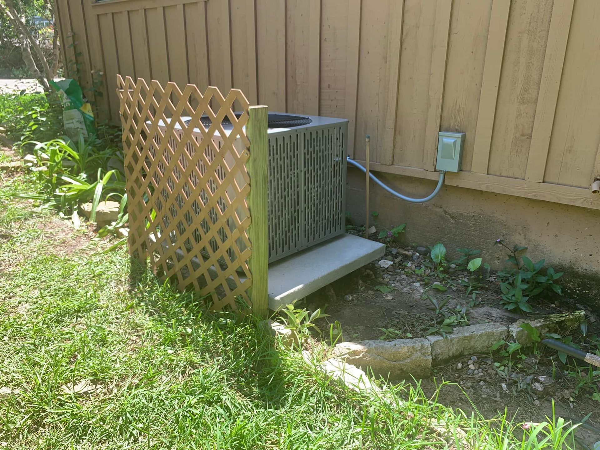 Outdoor HVAC unit beside a beige house, partially enclosed by a lattice fence and surrounded by grass.