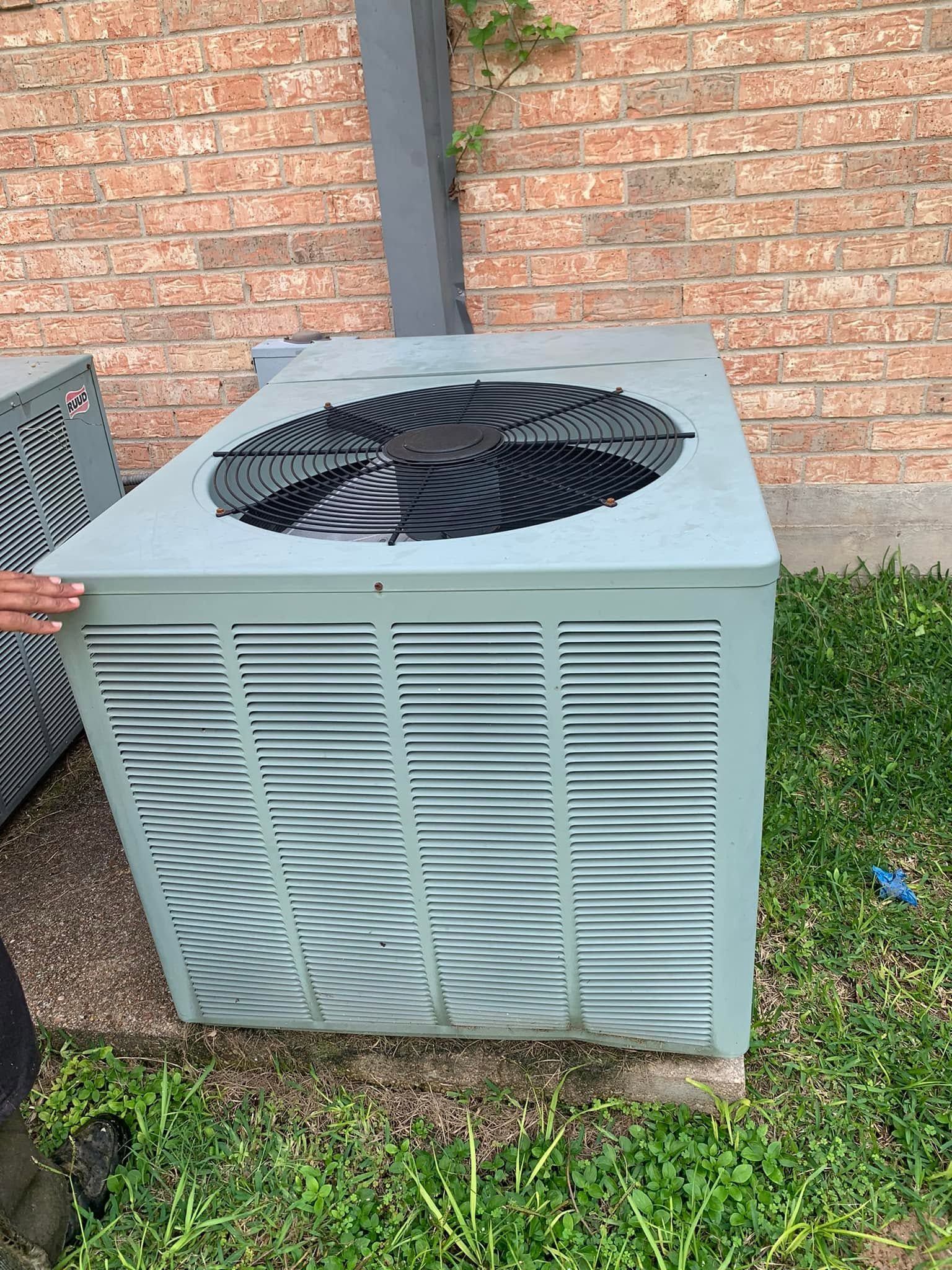 Outdoor HVAC unit with black fan on top against a brick wall and grass.