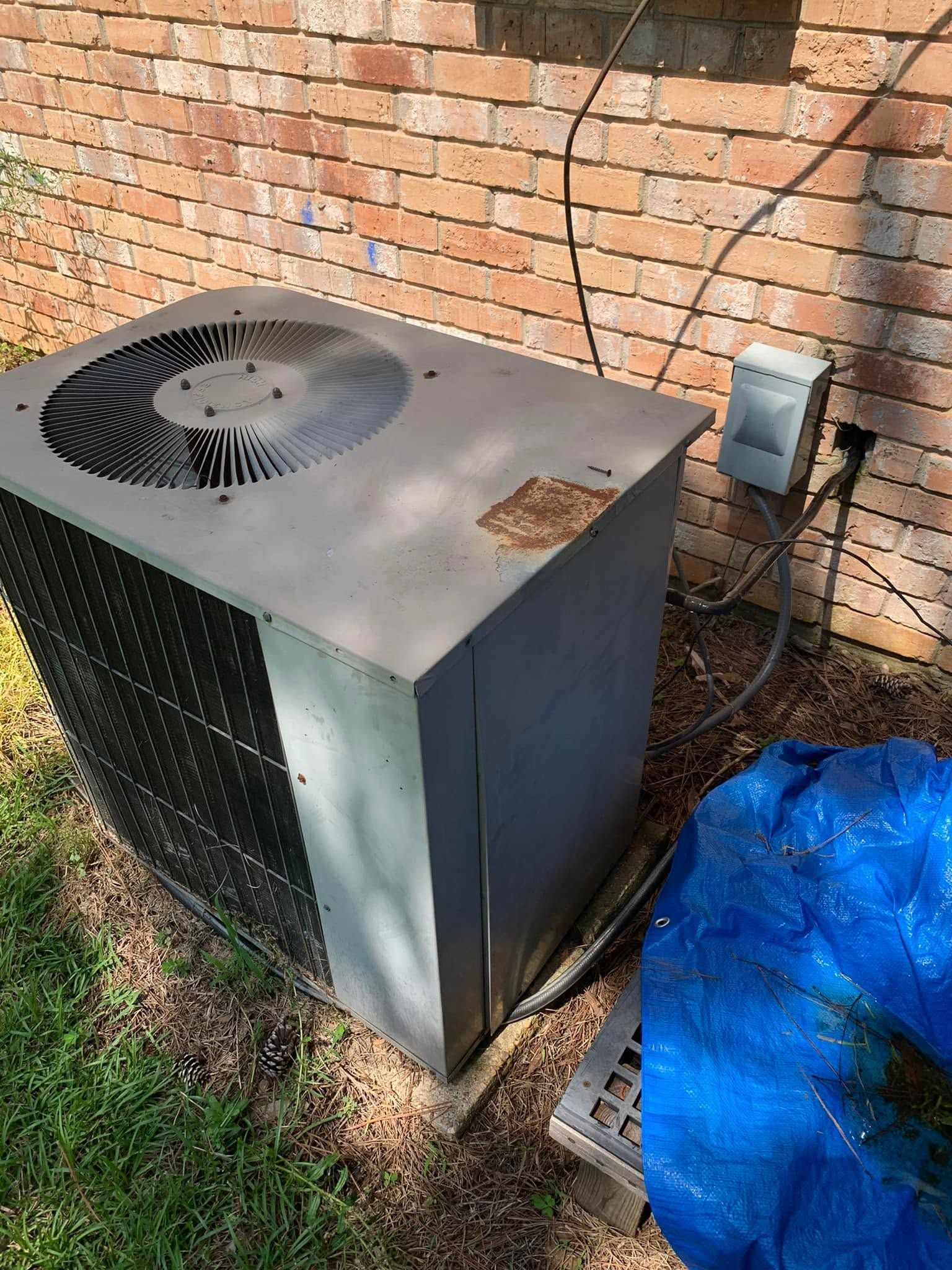 Outdoor HVAC unit beside brick wall, with electrical box, blue tarp, and fan visible on top.