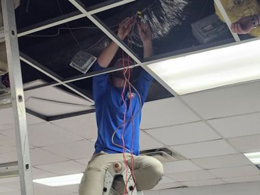 Worker in a blue shirt kneels in a dropped ceiling, handling wires during maintenance.