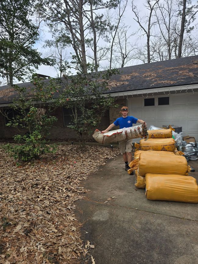 Person carrying a large sack beside stacked yellow bags in a yard with a white building and trees