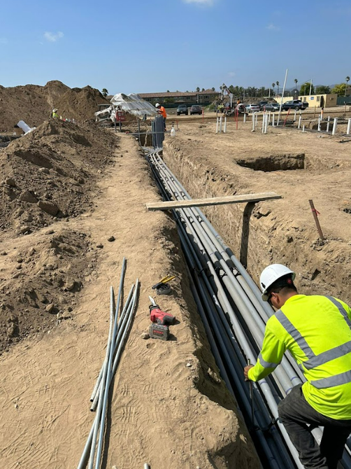 A worker in a yellow reflective shirt installs grey electrical conduits in a dirt construction trench.