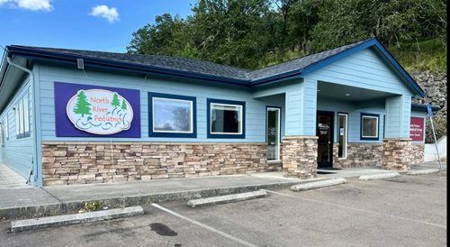 Small blue commercial building with stone base, parking lot, and forested hillside in the background