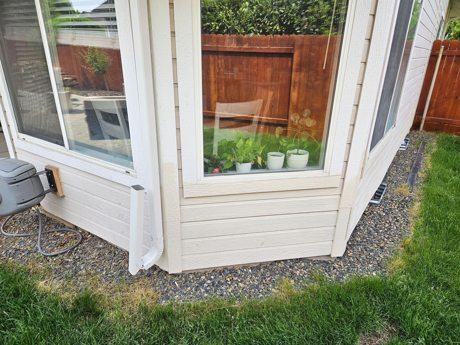 Beige house corner with windows, plants inside, gravel and grass below.