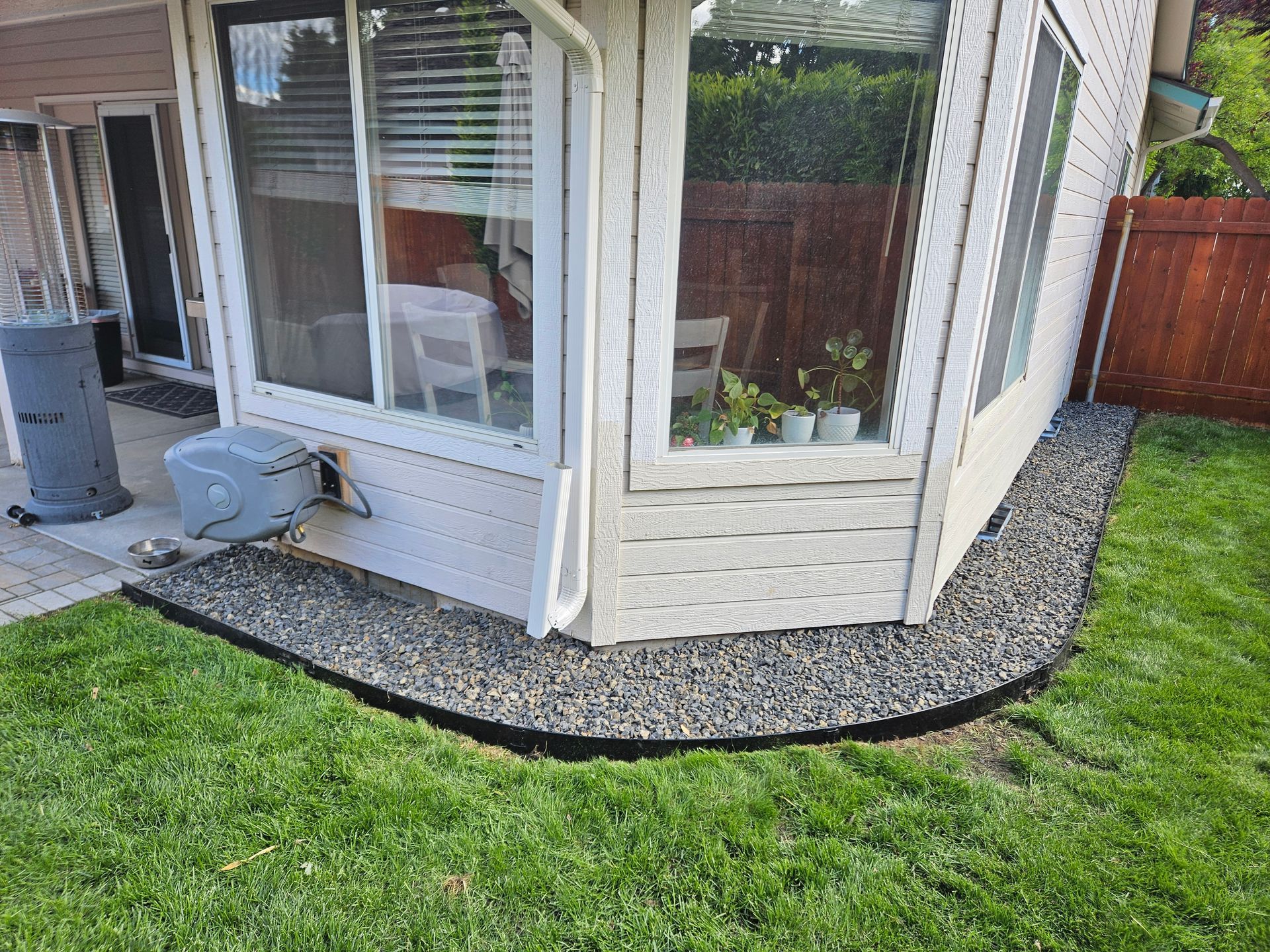 Corner of a house with a gravel border along the foundation. Green lawn, windows, and a patio heater are also visible.