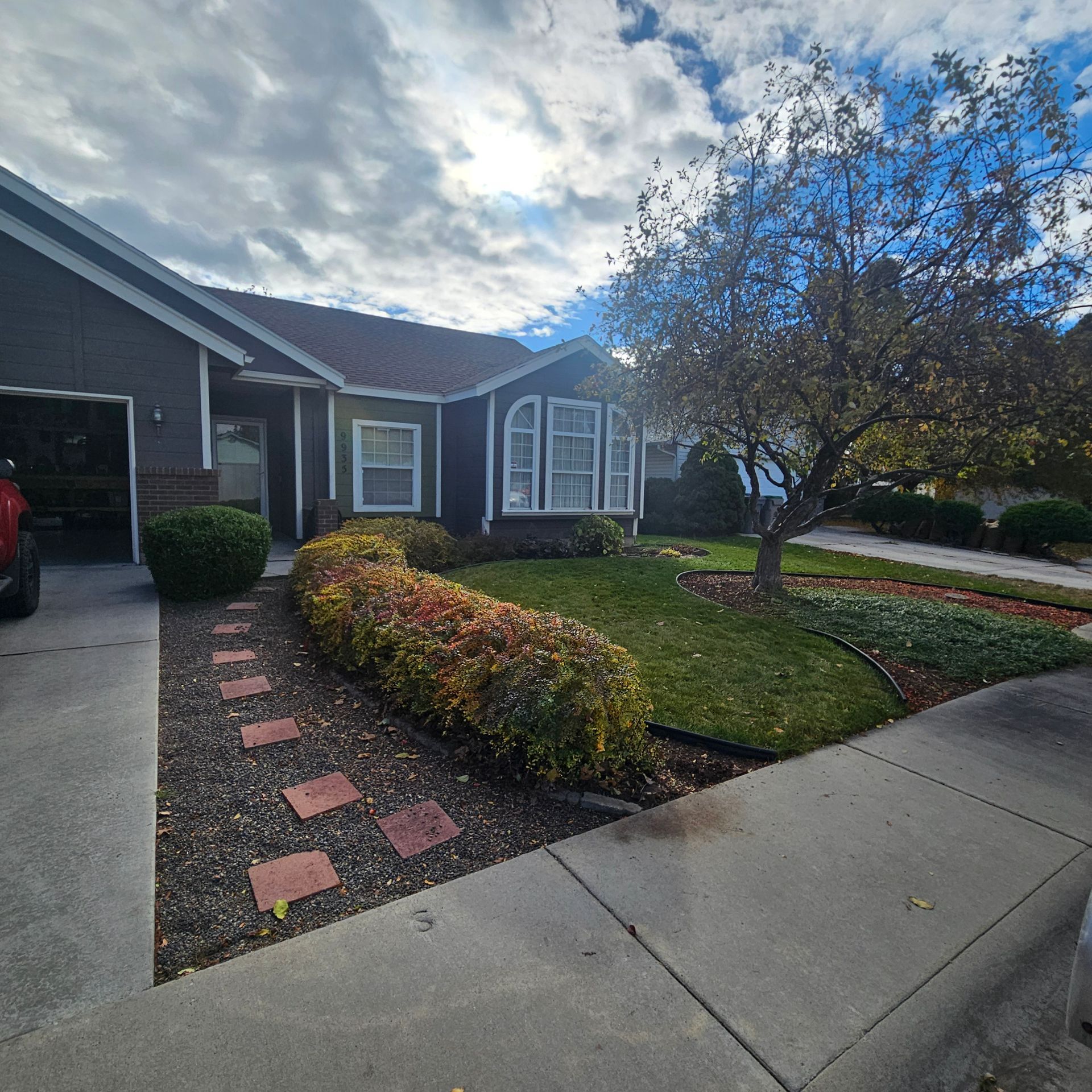 A house with a dark facade, surrounded by a landscaped yard with a brick pathway and a tree under a cloudy sky.